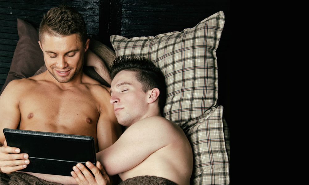 Gay couple lying together on a bed as they communicate sex chats with other gay people on their ipad.